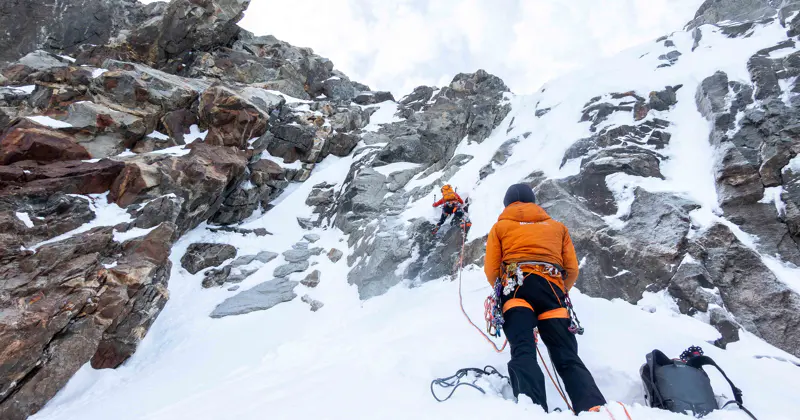 Twee klimmers in oranje jassen beklimmen een steile, besneeuwde en rotsachtige bergwand, aan elkaar vastgebonden met touwen. De grond en delen van de rotsen zijn bedekt met sneeuw. De klimuitrusting ligt vlakbij.